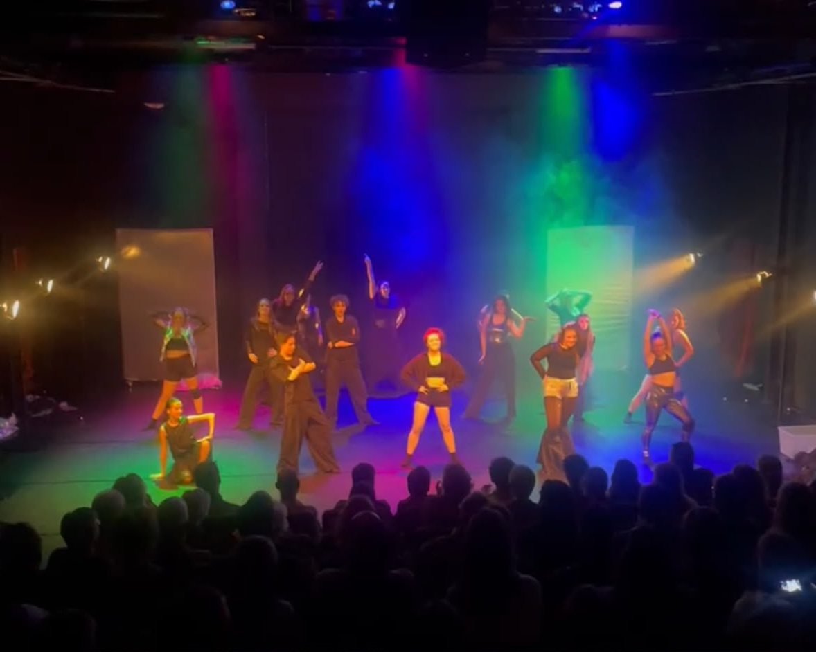 Dansers op het podium in kleurrijk paars en groen licht, met publiek op de voorgrond